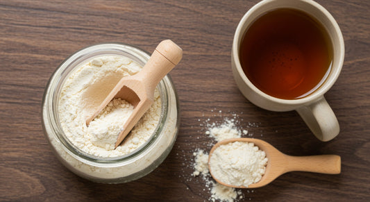 Glass jar of collagen peptide powder with a wooden scoop next to a ceramic mug of tea on a wooden surface