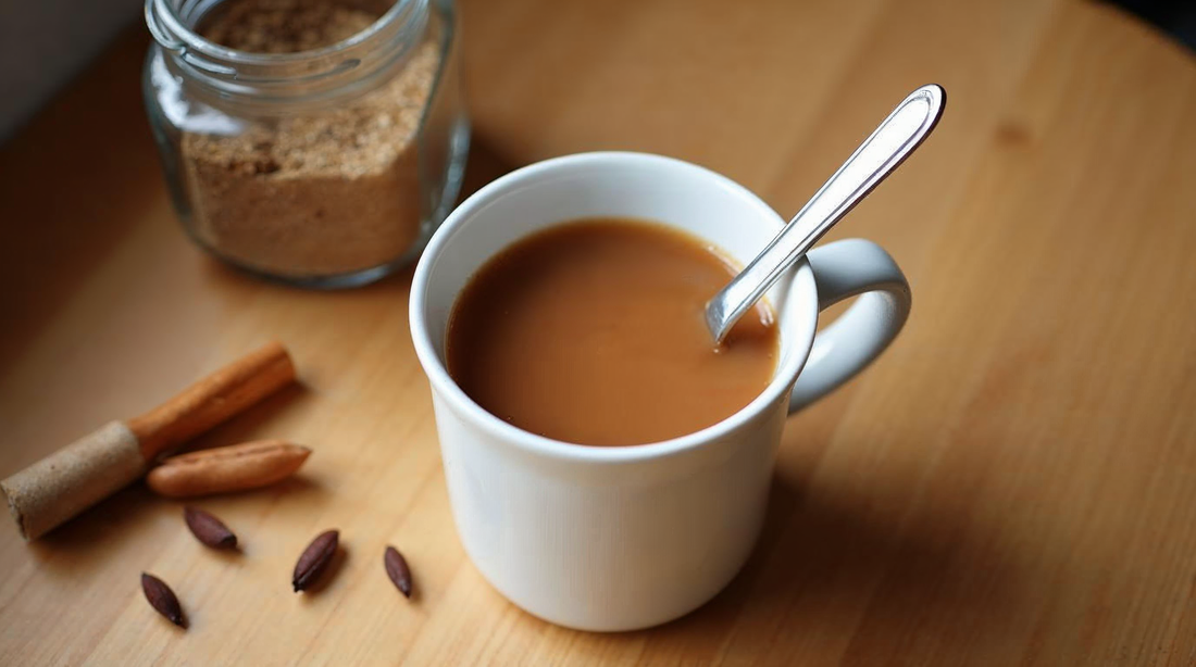 White ceramic mug of easy chai tea with a spoon, glass jar of milled chai blend in the background, cinnamon stick and cardamom pods on a wooden counter